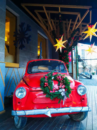A red car with Christmas decorations and decorated green wreath is standing on a street on wooden floor under yellow star shape Christmas lights. Festive New Year concept.の写真素材