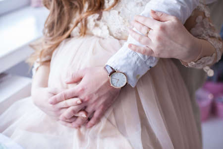 Close up of pregnant woman in beautiful pink dress holding her husbend hand and touching her belly. Happy pregnancy concept. Couple waiting for their child. Family love and care.の写真素材