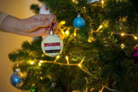 Close up of girls hand holding a Christmas ball for a fir tree with the flag of Egypt. New year in...