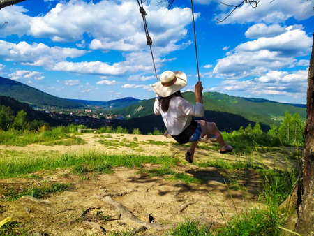 Back of girl in a straw hat, black and white blouse and blue shorts sitting on a swing with the mountain view. Calm and quiet wanderlust concept moment when person feels happiness and life.の写真素材