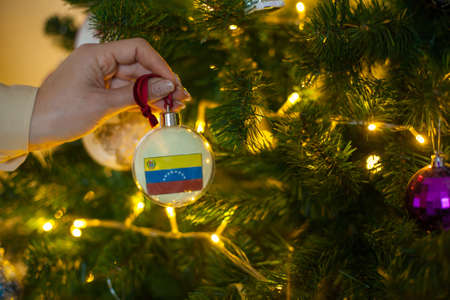 Close up of girls hand holding a Christmas ball for a fir tree with the flag of Venezuela. New year in Venezuela. Christmas holiday greeting card with copy space. Decorating home for celebration.の写真素材
