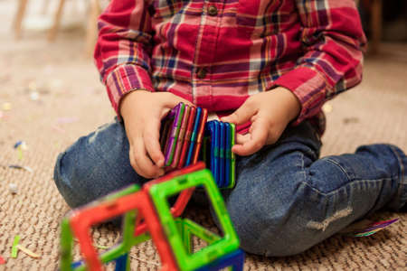 Cropped little boy sitting on the carpet and playing with a magnetic constructor. Close up of childs...