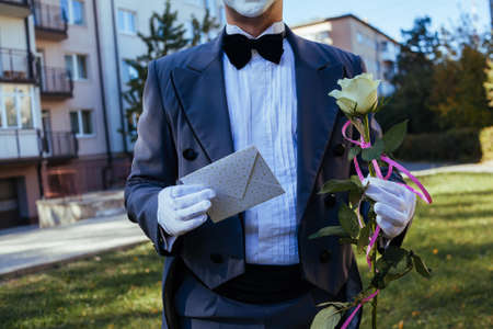 A man with hands in white gloves holds an envelope and white rose with pink ribbon. Cropped guy in a...