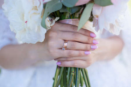 Hands of bride holding wedding bouquet. Close up of Bridal hands with elegant pink manicure with golden rings on the finger holding bunch of flowers on the wedding dress background.の写真素材