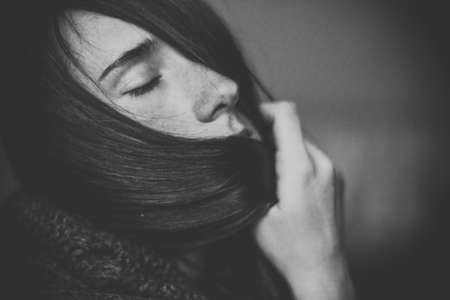 Black and white portrait of beautiful young girl with dark hair and closed eyes in black knitted sweater. Pretty woman without makeup at home. Atmospheric moody portrait.の写真素材