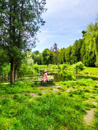 Back of girl in a straw hat and pink dress sitting on a bench with the lake view. Calm and quiet wanderlust concept moment when person feels happiness and life. Travelling in Ukraine in summer.の写真素材