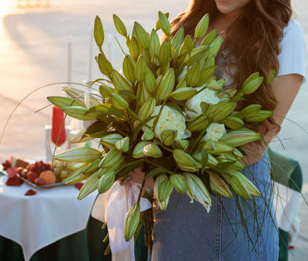 Woman in jeans dress holds a bouquet green lilies and white peonies in the background of table with fruits, wineglasses and candles. Girl with flowers. Proposal of marriage and small business conceptの写真素材