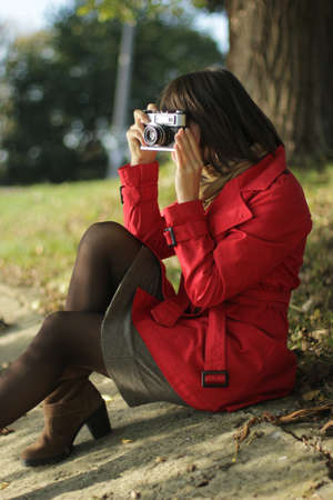 A happy smiling dark-haired girl in a red raincoat sits on the shore of the lake and holds the camera in her hands. Female photographer in coat and short dress taking pictures in the parkの写真素材