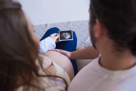 Pregnant woman holding photo of her unborn child while her husbands hand touching her belly. Happy pregnancy concept. Couple waiting for their child. Family love and care.の写真素材
