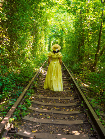 Scenic railway in the summer forest. Woman in yellow dress and straw hat walking in tunnel of love in Klevan, Ukraine. Railway surrounded by green arches. It is known romantic place for couples.の写真素材