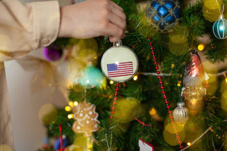 Close up of girls hand holding a Christmas ball for a fir tree with the flag of USA. New year in USA. Christmas holiday greeting card with copy space for text. Decorating home for celebration.の写真素材