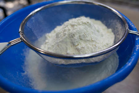 Sifting white flour through a sieve into blue bowl on table on wooden background. Flour for dough...