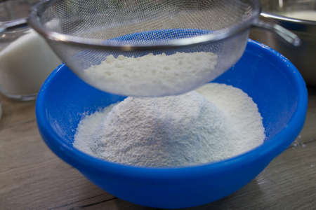 Sifting white flour through a sieve into blue bowl on table on wooden background. Flour for dough...