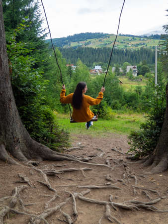 Back of girl with dark hair in a mustard sweater sitting on a swing with the mountain view. Calm and quiet wanderlust concept moment when person feels happiness and life. Alone travel in Ukraine.の写真素材