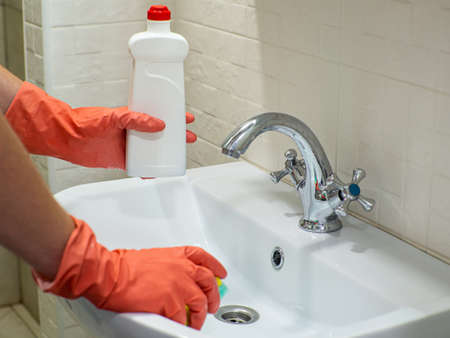 Cleaning bathroom sink and faucet with detergent in orange rubber gloves with green sponge. Housework, cleaning, hygiene home concept. Person hand holding a detergent in white bottle for cleaning sinkの写真素材