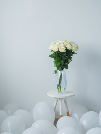 Beautiful bouquet of white blooming roses in glass vase on the white background. White flowers with ribbon stand on small round table with white balloons. Floral still life. Rose hybrid tea Avalanche.の写真素材