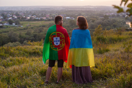 Couple covered in Ukraine and Portugal flags. Concept of friendship between Ukraine and Portuguese Republic. Love between two countries. Man and woman cover different flags on background of sunsetの写真素材