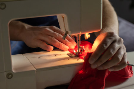 Hands of experienced worker in the handmade industry, sewing red fabric on a sewing machine, the production of Santa red hat. Homemade sewing. Working tailor. Christmas hat. Close up.の写真素材