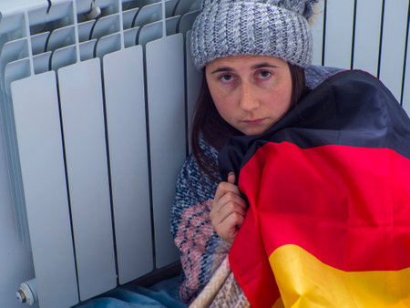 Woman sitting near the battery at home and trying to get warm covered in german flag. Warmly dressed woman is upset about the high price of heating. Expensive energy resources, energy crisis in Europeの写真素材