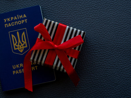 Small gift in a stripped black and white wrapper and with red ribbons and passport of Ukraine on the dark background. Travel for Ukrainians, a trip as a gift concept. Copy space for your text.の写真素材