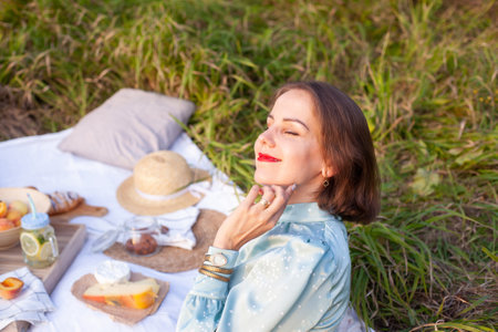 A girl in a long summer dress with short hair lying on a white blanket with fruits and pastries and enjoying the moment. Concept of having picnic in a city park during summer holidays or weekends.の写真素材