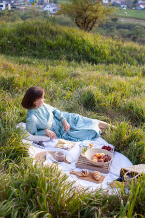 A girl in a long summer dress with short hair lying on a white blanket with fruits and pastries and enjoying the view. Concept of having picnic in a city park during summer holidays or weekends.の写真素材