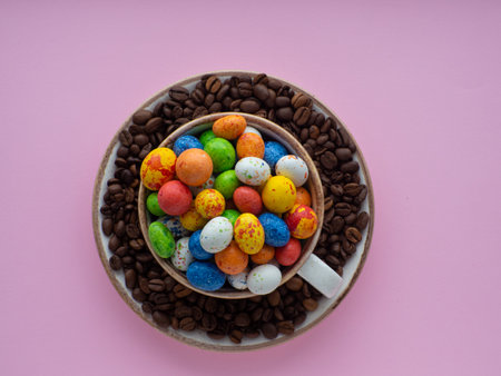 Flat lay composition with small chocolate easter eggs in cup surrounded by coffee roasted beans. Easter coffee concept. Mug full of colorful eggs on a plate with coffee beans on a pink background.の写真素材