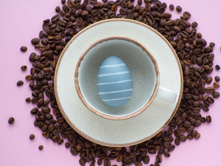 Flat lay composition with easter egg in cup surrounded by coffee roasted beans. Easter coffee concept. Mug full of gray egg on a plate on a pink background with a lot of coffee beans and copy space.の写真素材