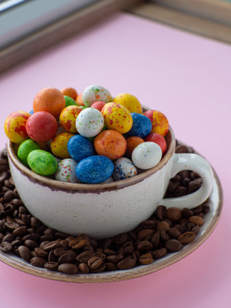 Composition with small chocolate easter eggs in cup surrounded by coffee roasted beans. Easter coffee concept. Mug full of colorful eggs on a plate with coffee beans on a pink background.の写真素材