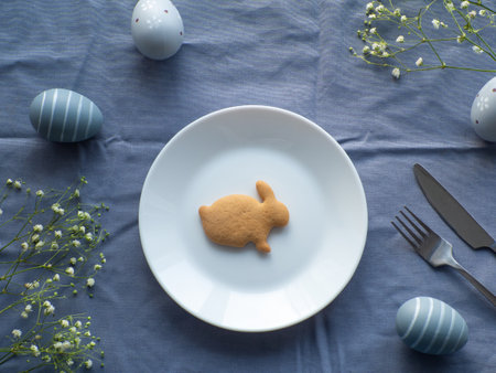 Festive Easter table setting with flowers, eggs and bunny shape cookie on blue background, flat lay. Elegance tablescapes. Easter celebration. Delicate Easter composition. View from above.の写真素材