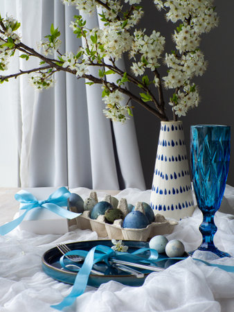 Festive Easter table setting with flowers, painted eggs and gift box on white background. Elegance pastel and blue tablescapes. Easter celebration. Delicate Easter composition. View from side.の写真素材
