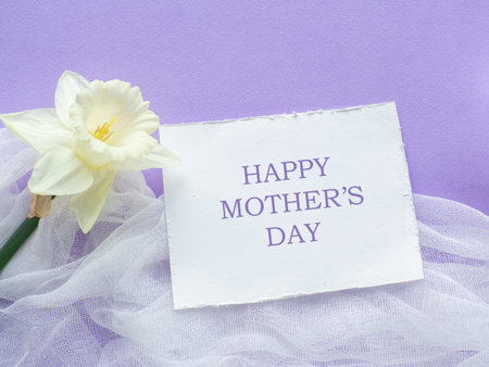 Daffodils flower on lilac background with Happy mothers day on white card. Mothers Day greeting card. Sheet of paper with text and narcissus on purple background. Congratulations note flat lay.の写真素材