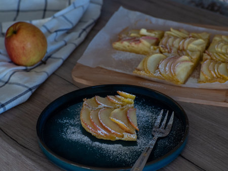 Apple vanilla squares. Homemade apple pie topped with slices of apples. Serving of delicious fruit vanilla cake on a dark plate with fork. Thin slices of apples are stuffed tightly and powdered sugar.の写真素材