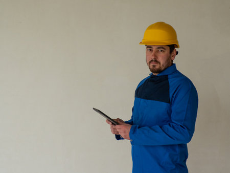 Portrait of worker or engineer in blue work clothes and yellow safety helmet with space for text. A man at a construction site holds a tablet in his hands and looks at the camera.の写真素材