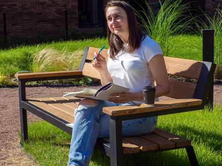 Pleased brunette woman sitting on bench and reading book in park. Education, school and people concept. Female student studying on bench at campus. Girl sitting with book, coffee cup and writing.の写真素材