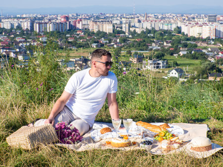 A stylish man in light clothes and black sunglasses sits on the blanket and waiting for his date. Having a picnic with summer fruits, pastries and drinks. Date with cityscape. Copy spaceの写真素材
