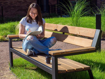 Pleased brunette woman sitting on bench and reading book in park. Education, school and people concept. Female student studying on bench at campus. Girl sitting with book, coffee cup and writing.の写真素材