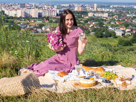 A woman in a pink dress and with dark hair sitting on a beige blanket holding glass and drinking white wine. Concept of having picnic in a city park during summer holidays or weekends. Pink flowers.の写真素材