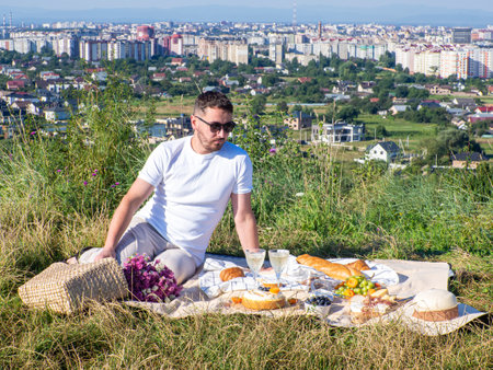 A stylish man in light clothes and black sunglasses sits on the blanket and waiting for his date. Having a picnic with summer fruits, pastries and drinks. Date with cityscape. Copy spaceの写真素材