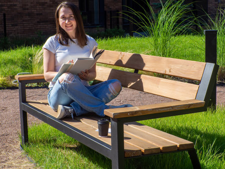 Pleased brunette woman sitting on bench and reading book in park. Education, school and people concept. Female student studying on bench at campus. Girl sitting with book, coffee cup and writing.の写真素材