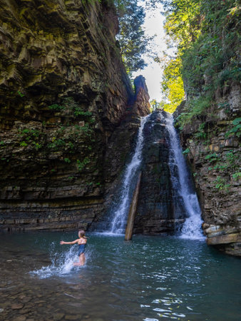 Small girl swimming in the mountain river. Rear view of girl walking under the waterfall. Child is...