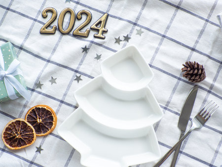 Empty christmas tree shape white plate with 2024 on towel background. New year porcelain plate among the cones of a fir tree, decorative stars, dried orange and gift box. Top view flat lay, copy spaceの写真素材