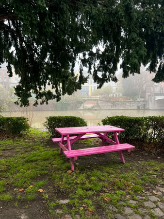 A picnic table made of wood is placed beneath a tree in a park, surrounded by grass and embraced by...