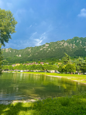 Stunning landscape featuring a calm lake reflecting the cloudy sky. Majestic mountains rise in the distance, framed by lush greenery, creating a tranquil and picturesque scene. Lake Idro, Crone, Italyの写真素材