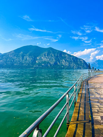 A stunning landscape featuring a calm Iseo lake reflecting the blue sky, Italy. Majestic mountains rise in the distance, framed by lush greenery, creating a tranquil and picturesque scene.の写真素材