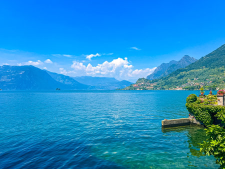 A stunning landscape featuring a calm Iseo lake reflecting the blue sky, Italy. Majestic mountains rise in the distance, framed by lush greenery, creating a tranquil and picturesque scene.の写真素材
