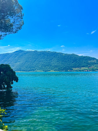 A stunning landscape featuring a calm Iseo lake reflecting the blue sky, Italy. Majestic mountains rise in the distance, framed by lush greenery, creating a tranquil and picturesque scene.の写真素材