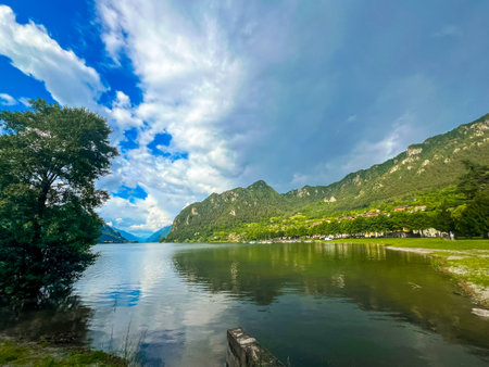 Stunning landscape featuring a calm lake reflecting the cloudy sky. Majestic mountains rise in the distance, framed by lush greenery, creating a tranquil and picturesque scene. Lake Idro, Crone, Italyの写真素材
