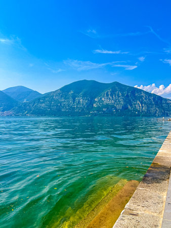 A stunning landscape featuring a calm Iseo lake reflecting the blue sky, Italy. Majestic mountains rise in the distance, framed by lush greenery, creating a tranquil and picturesque scene.の写真素材