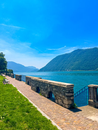 A stunning landscape featuring a calm Iseo lake reflecting the blue sky, Italy. Majestic mountains rise in the distance, framed by lush greenery, creating a tranquil and picturesque scene.の写真素材